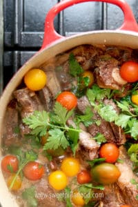overhead shot of quarter of red pot on black stove showing short ribs in broth with multicolored cherry tomatoes and fresh 